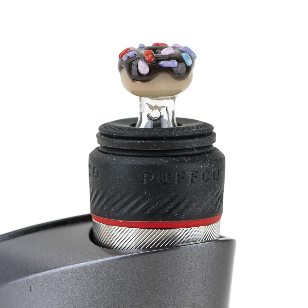 A close-up shows the Donut PuffCo Peak Pro Glass Ball Cap, a translucent, spherical cap with a colorful, donut-like design. A close-up shows the Donut PuffCo Peak Pro Glass Ball Cap, a translucent, spherical cap with a colorful, donut-like design.