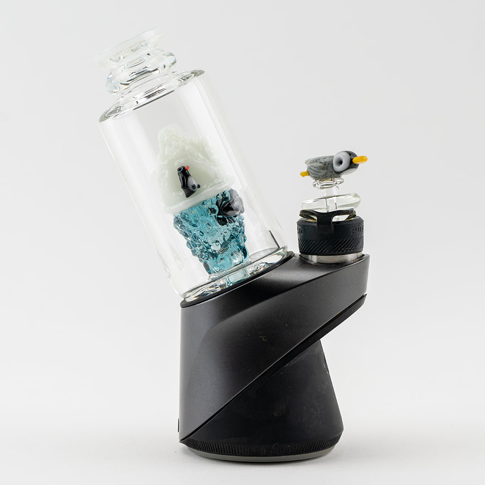 A side view of the Avenge the Arctic PuffCo Peak & Peak Pro Glass Attachment, displaying a translucent blue glass piece atop a black base. A side view of the Avenge the Arctic PuffCo Peak & Peak Pro Glass Attachment, displaying a translucent blue glass piece atop a black base.