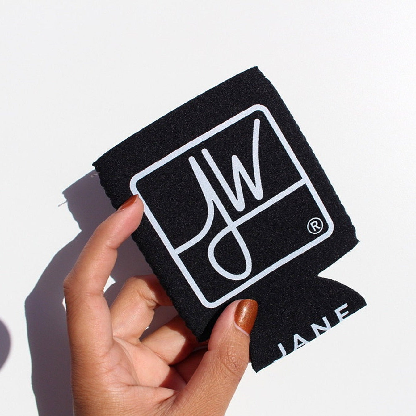 A hand holds the Jane West Koozie with Custom Pocket, featuring a sleek, dark-colored design in a bright, angled studio shot. A hand holds the Jane West Koozie with Custom Pocket, featuring a sleek, dark-colored design in a bright, angled studio shot.