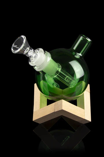A front view of The Orb Bubbler, a green glass piece displayed on a light wood stand against a black background. A front view of The Orb Bubbler, a green glass piece displayed on a light wood stand against a black background.