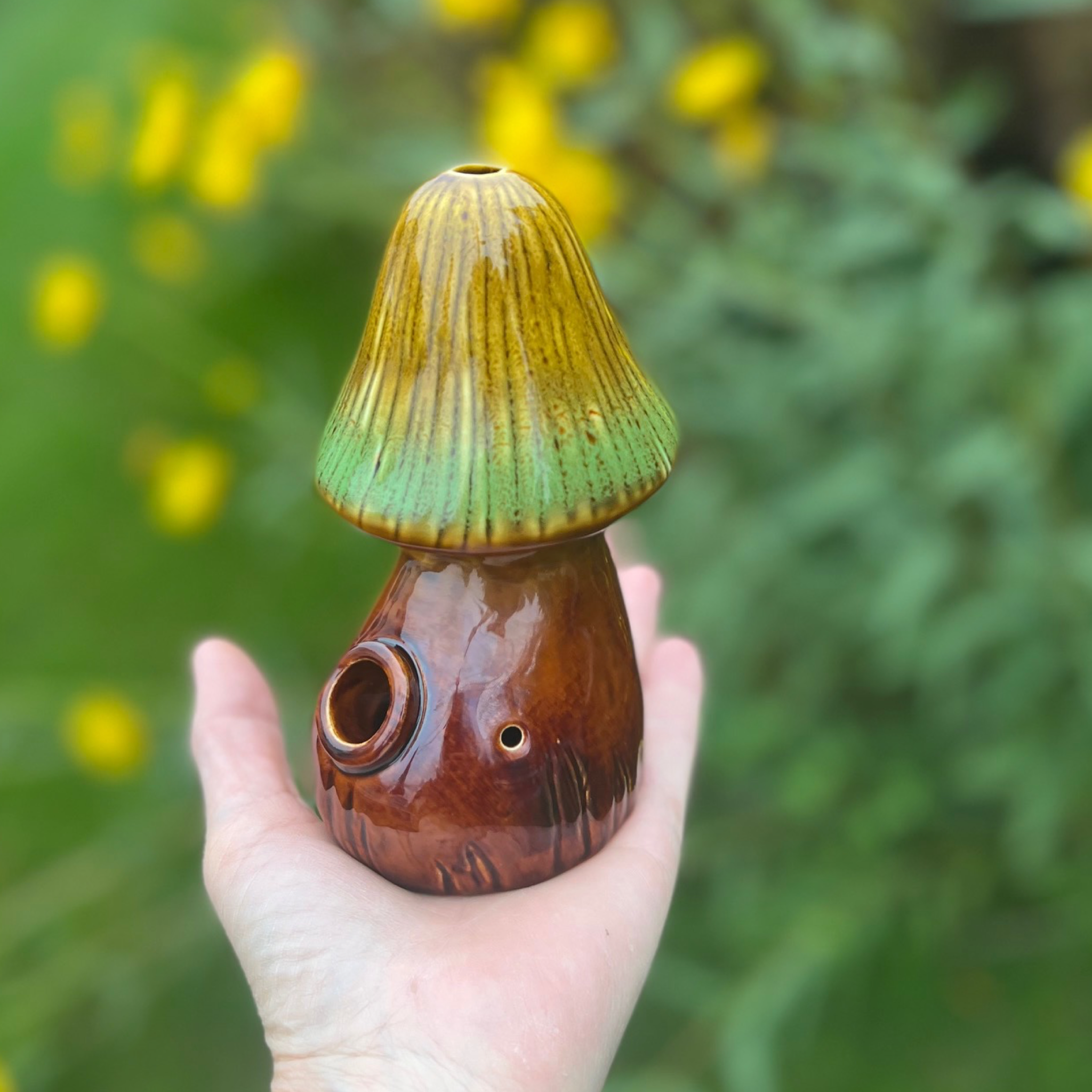 A hand holds the Mushroom Pipe, showcasing its earthy-toned, sculpted form against a blurred green background. A hand holds the Mushroom Pipe, showcasing its earthy-toned, sculpted form against a blurred green background.