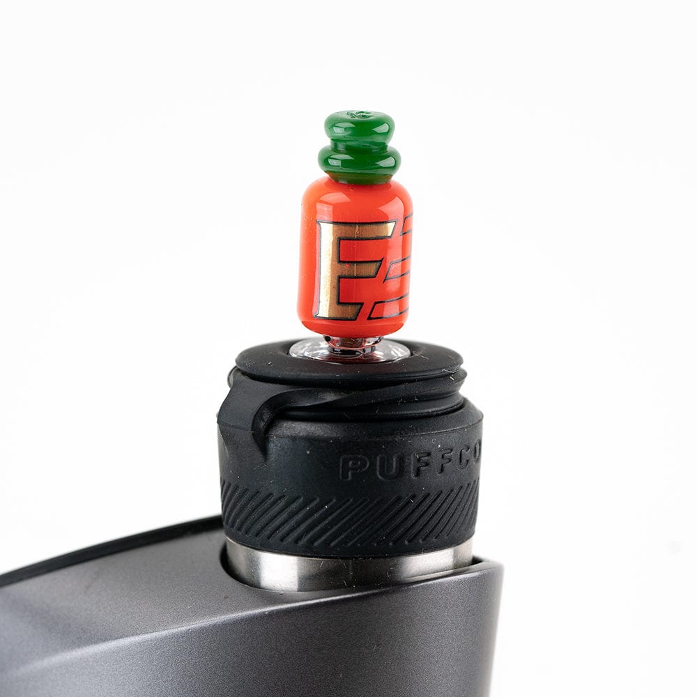 A close-up shows the Sriracha Bottle Puffco Peak Pro carb cap resting on a black surface, mimicking the iconic hot sauce bottle. A close-up shows the Sriracha Bottle Puffco Peak Pro carb cap resting on a black surface, mimicking the iconic hot sauce bottle.