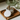 The Grinder rests open on a wooden surface, revealing ground plant material inside its metallic, circular frame.